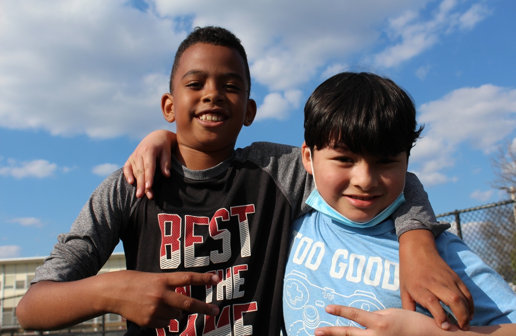 Two young boys pose together with their arms around each other's shoulders on a sunny field. 