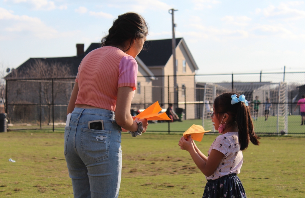 Photo of Brown student teaching a young elementary school girl how to throw a paper airplane. 