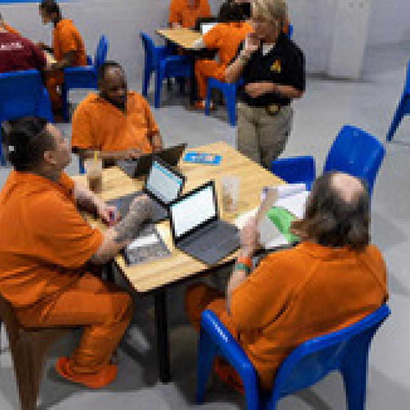 Photo of incarcerated individuals sitting at a table working on computers.