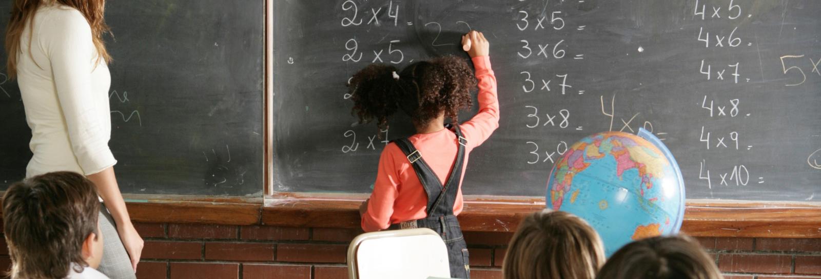 a young girl solves simple math problems on the black board while teacher and classmates watch