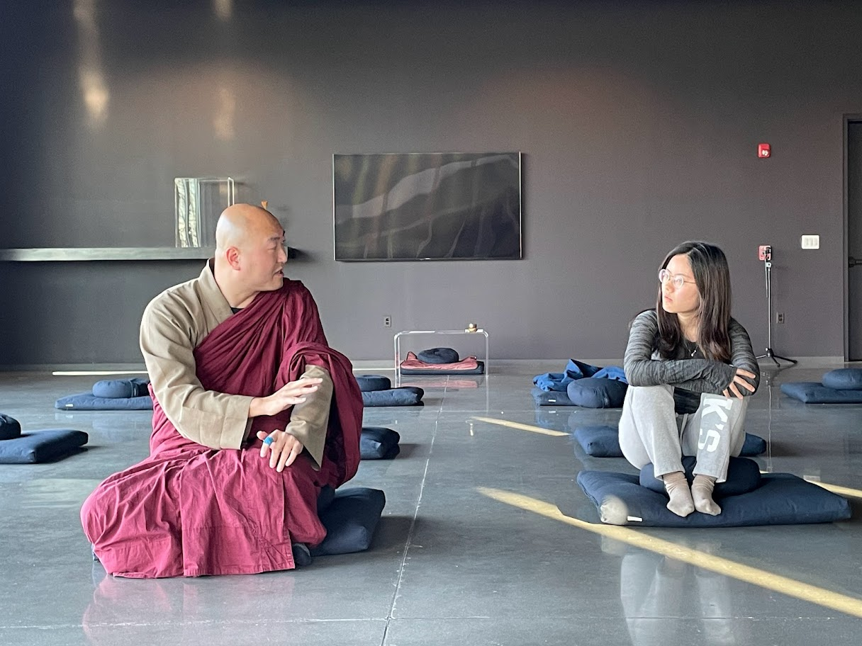 student sits on a meditation cushion next to a monk