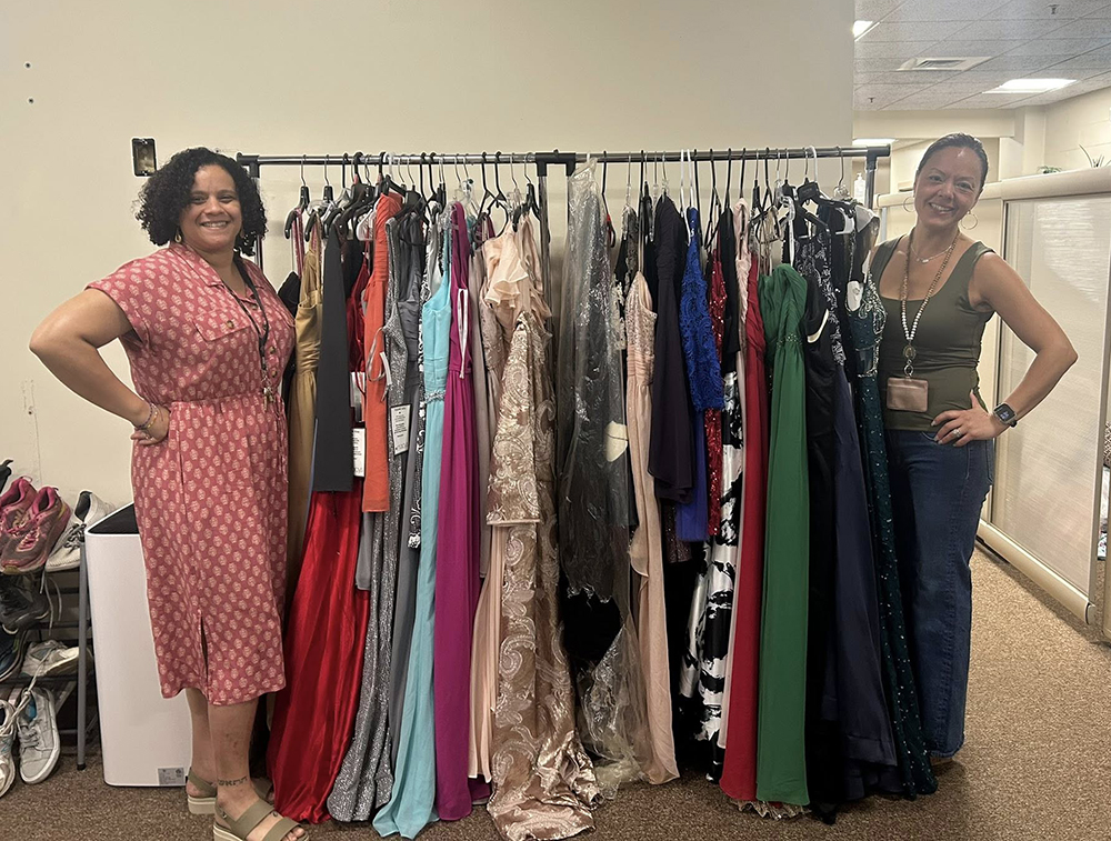 Two school administrators stand and smile next to a rack of donated prom dresses. 