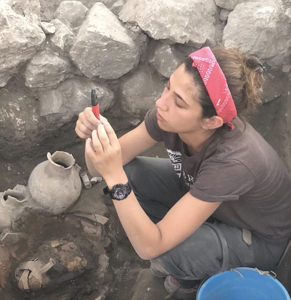 Rachel Kalisher examining an artifact while kneeling at an excavation site