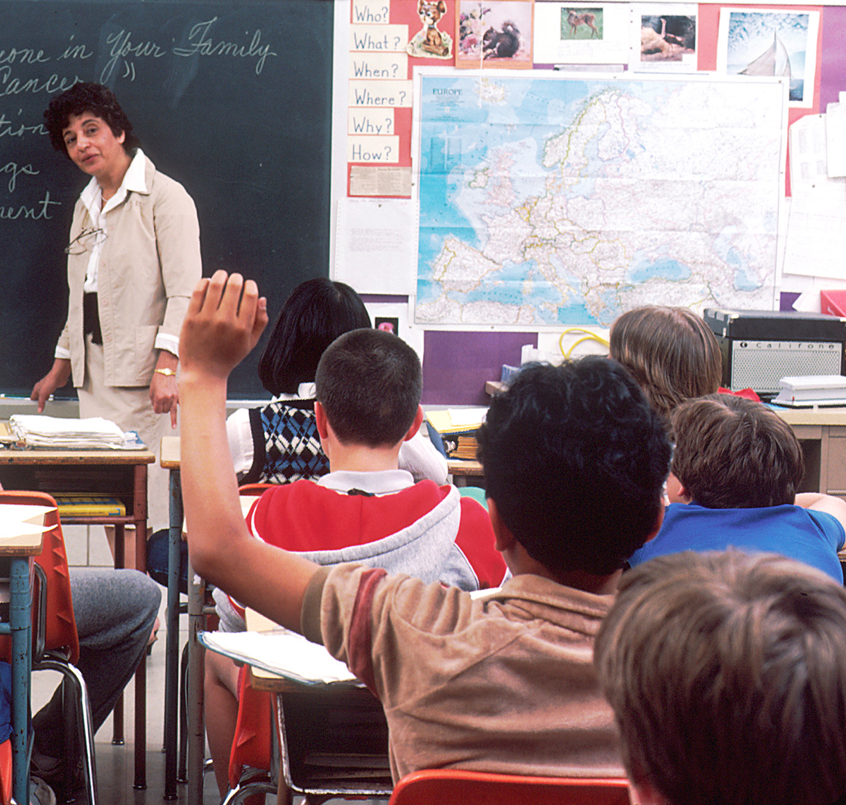 Teacher in front of classroom at blackboard, student raising hand