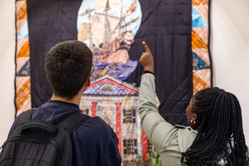 two students pointing at a tapestry depicting a slave ship and a colonial building