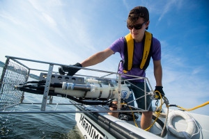 Brown student holds giant piece of technology over the side of a boat with ocean beneath