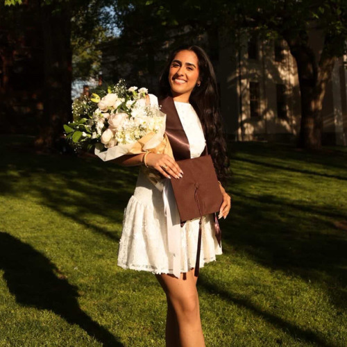 A recent Brown grad in a dress and sash stands on the green holding flowers.