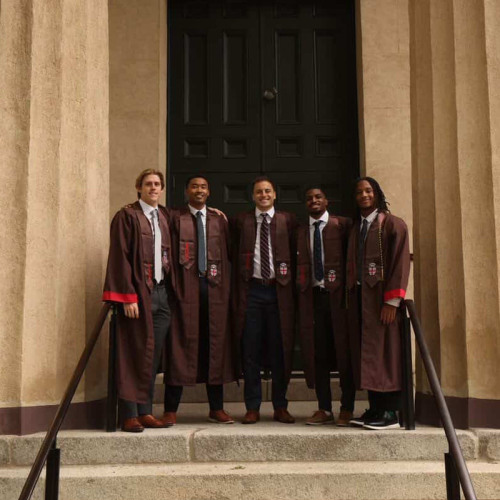 Five Brown Grads in regalia pose between columns outside an academic building.