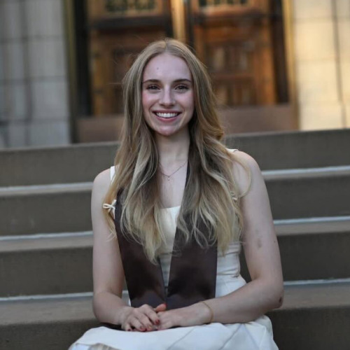 A Brown grad in regalia sits on the steps of an academic building.