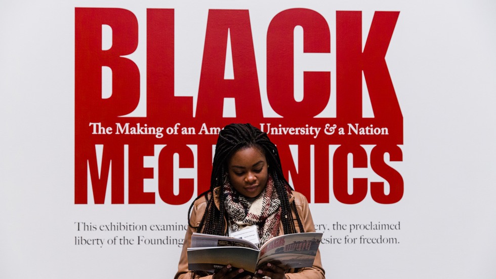 A young woman stands in front of the Black Mechanics exhibit poster