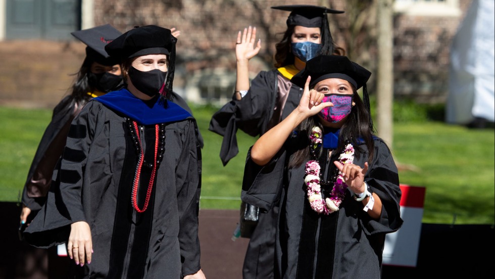 hooded and masked students waving at a camera