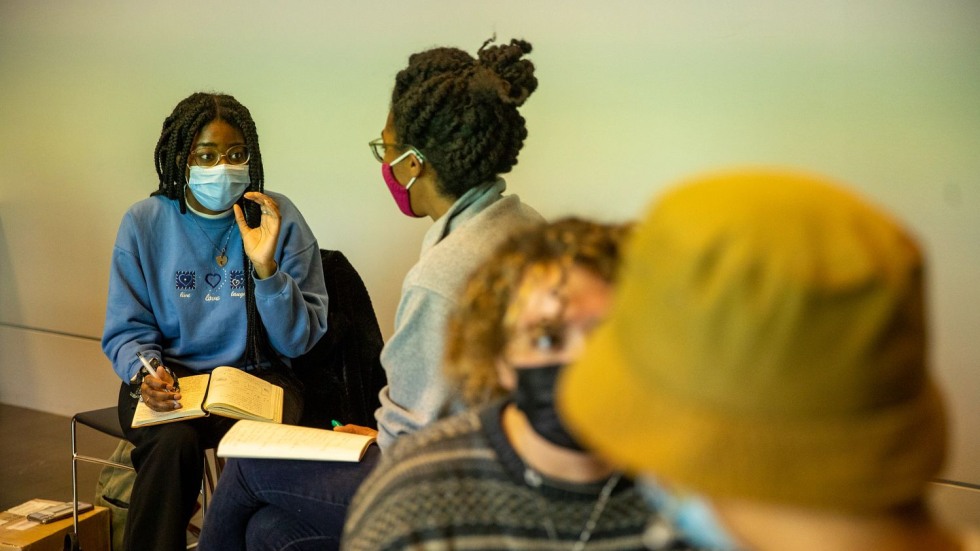 Two masked students in a discussion with notebooks on their laps