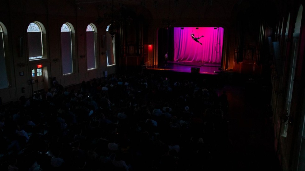 man hanging from a trapeze on a stage with darkened audience looking on