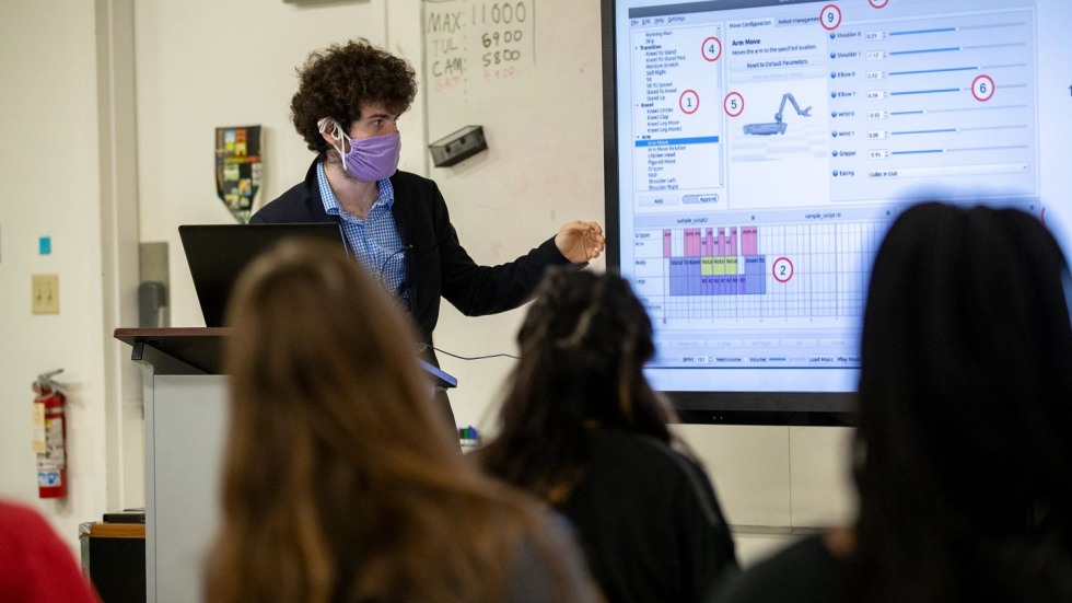 masked person gesturing toward a large TV monitor in front of a class 