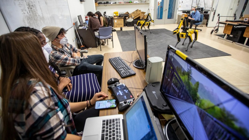 three students sitting at a desk with two computers, looking at a robot