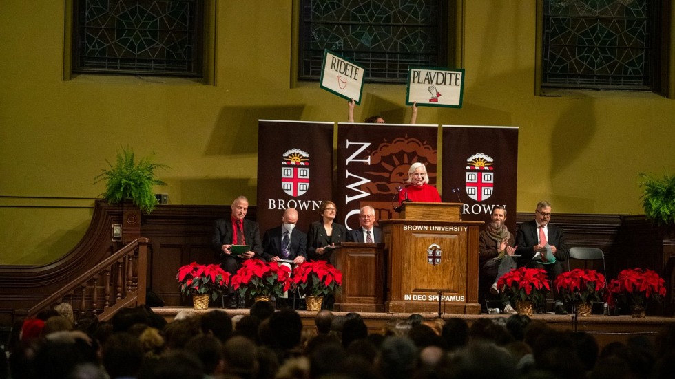 woman standing at a podium with "plaudite" and "ridete" signs behind her