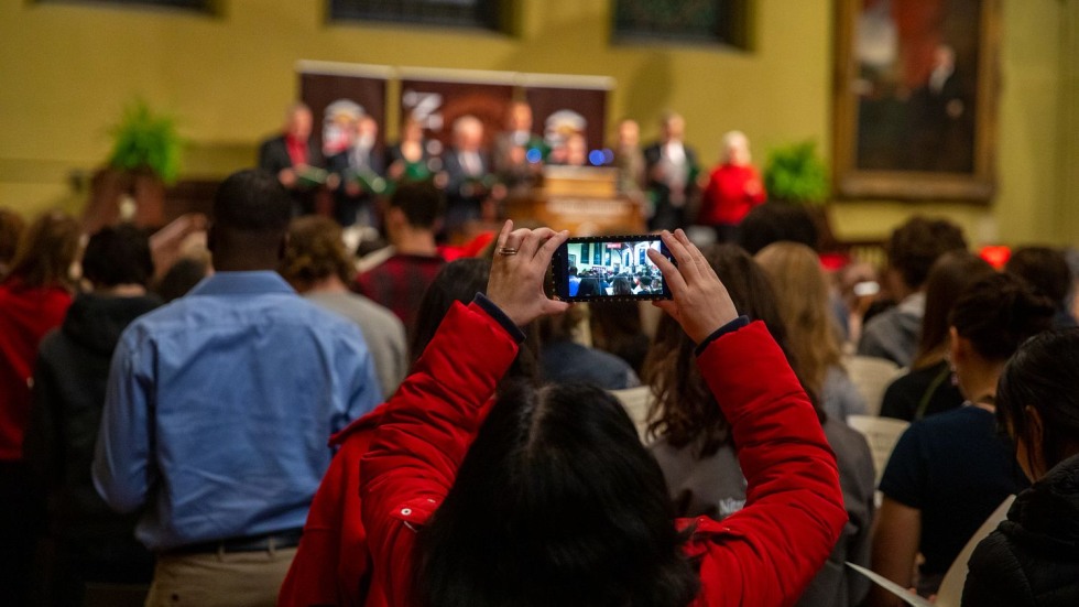 woman taking a cell phone picture of people standing at a podium