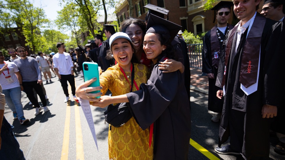 two students in caps and gowns embracing a woman in a yellow dress