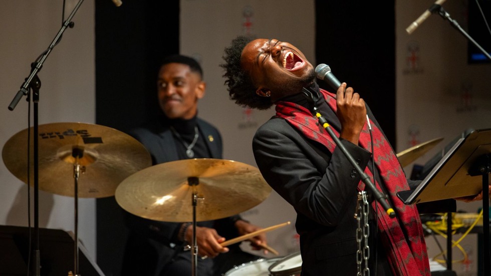man singing into a micrphone on a stage with a percussionist in the background