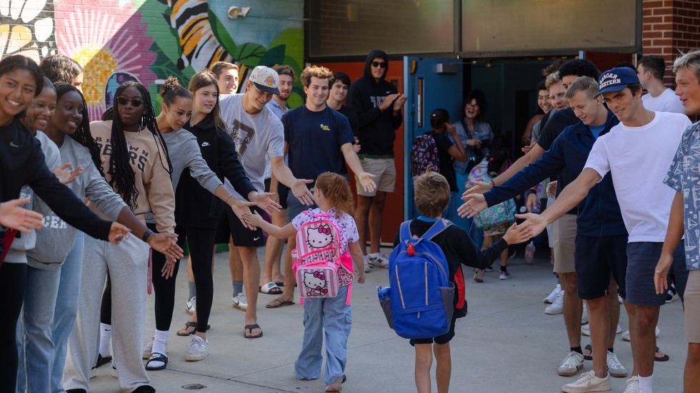Brown student-athletes outstretch arms to offer high-fives to young students as they enter the school building.