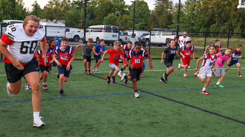 Brown football players race across a field with young kids as part of football clinic. 