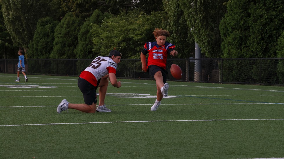 Young kids practice kicking a football, guided by Brown football player.