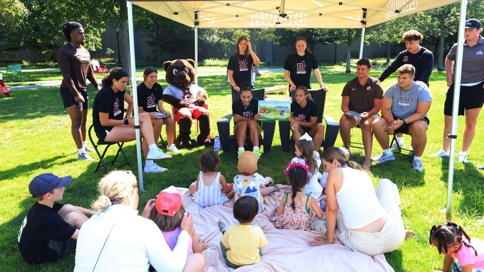 Brown student-athletes hold up books and read aloud to families sitting on blankets.