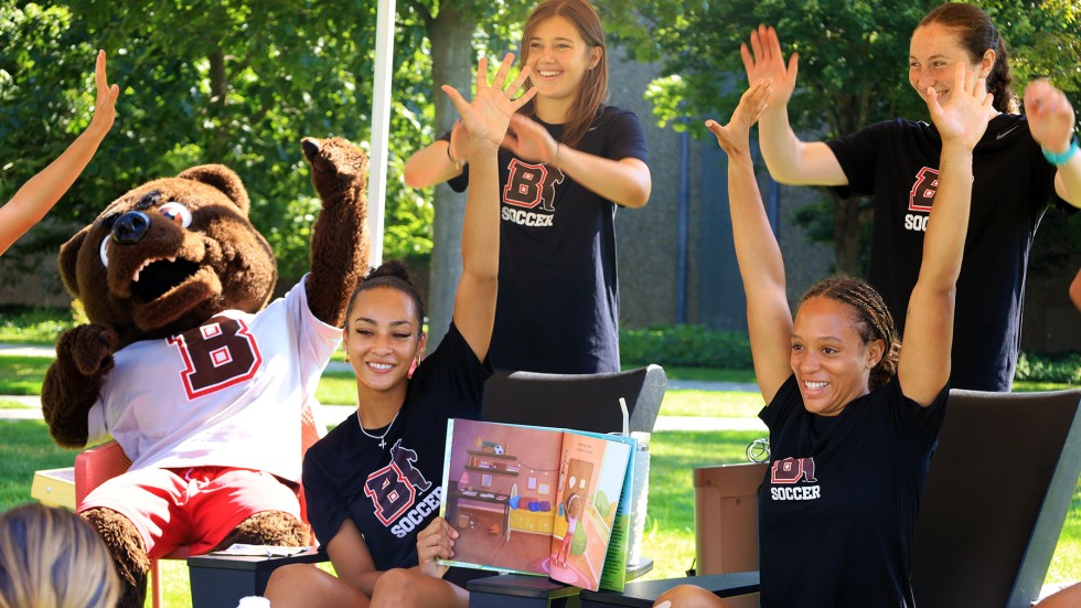 Brown student-athletes hold up books as they read to a group of kids sitting on blankets on a green space on campus.