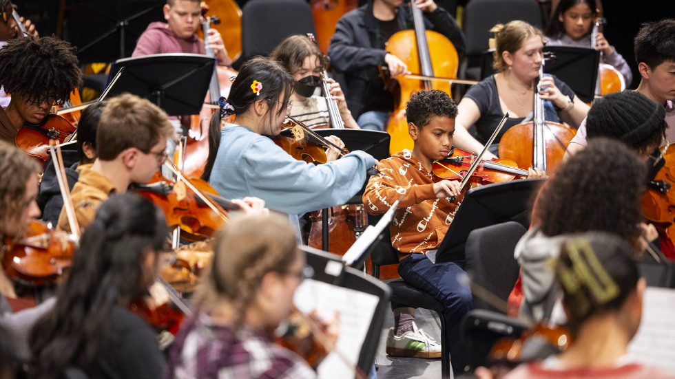 Providence K-12 student sit alongside the Brown University Orchestra with their instruments readying to play a song together.
