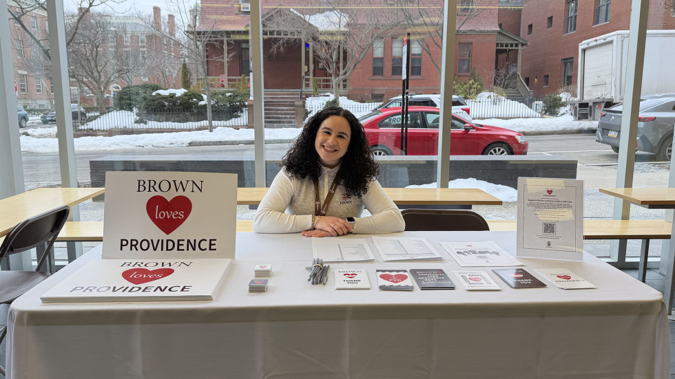 A Brown staff member smiles behind a table with Brown Loves Providence cards, posters and stickers. 