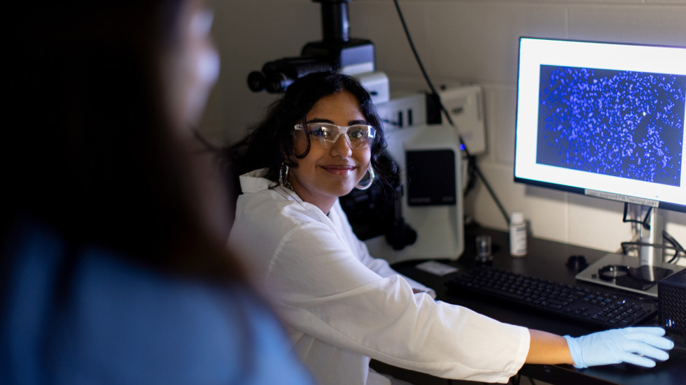 Rishika Kartik smiles at a labmate while looking at dyed cells under a microscope