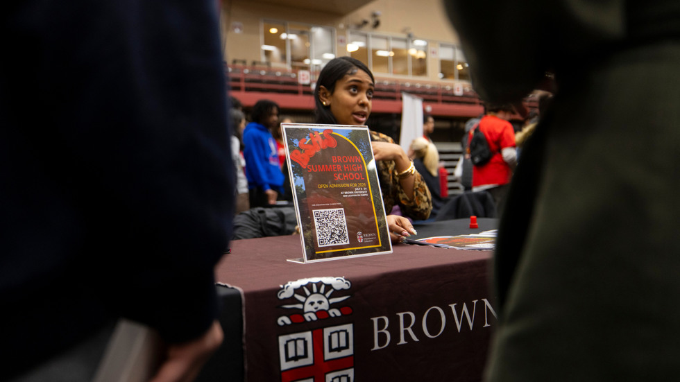 A Brown staff member sits behind a table at the college fair talking to local students about Brown Summer High School.