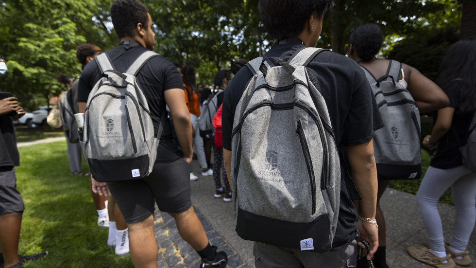 Students stand on a walkway on Brown's campus with gray backpacks on, branded with Brown Collegiate Scholars