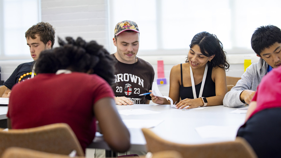 A instructor smiles with a student at a table, working on a group assignment together.
