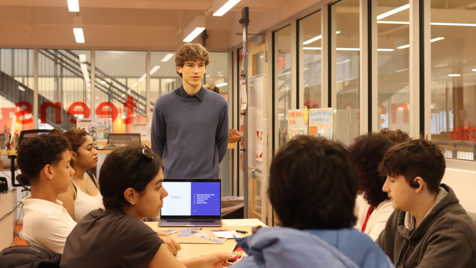 Brown student Justin Dunn stands at the front of a table at the library where students taking his English sit and look toward him.