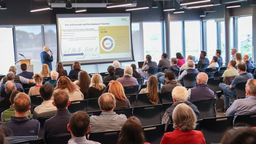 A group of nearly 100 participants sit in a room listening to a pitch presentation. 
