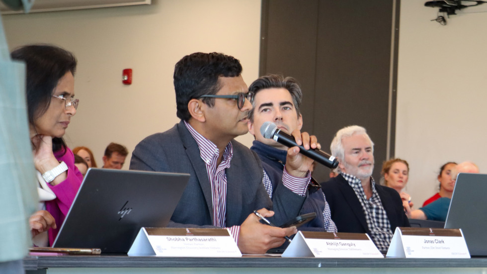 A group of professionals sit among a row of tables asking questions of presenters.