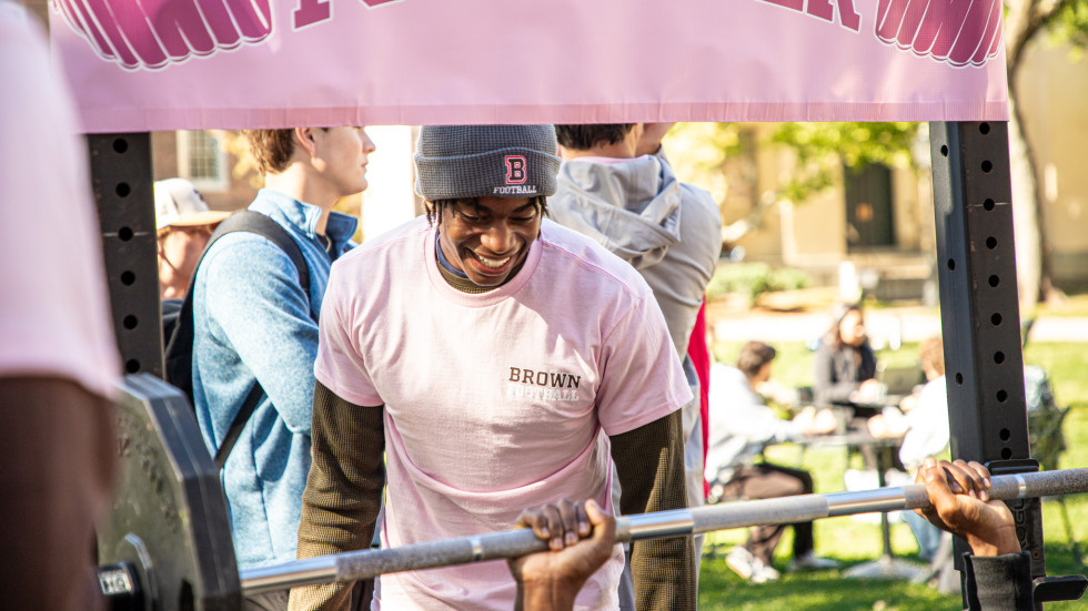 Brown football players spot participants who are pushing heavy weights as part of bench press exercise 