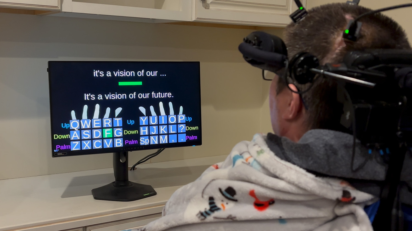 A BrainGate participant wearing a head-mounted sensor system, typing on a virtual keyboard displayed on a monitor
