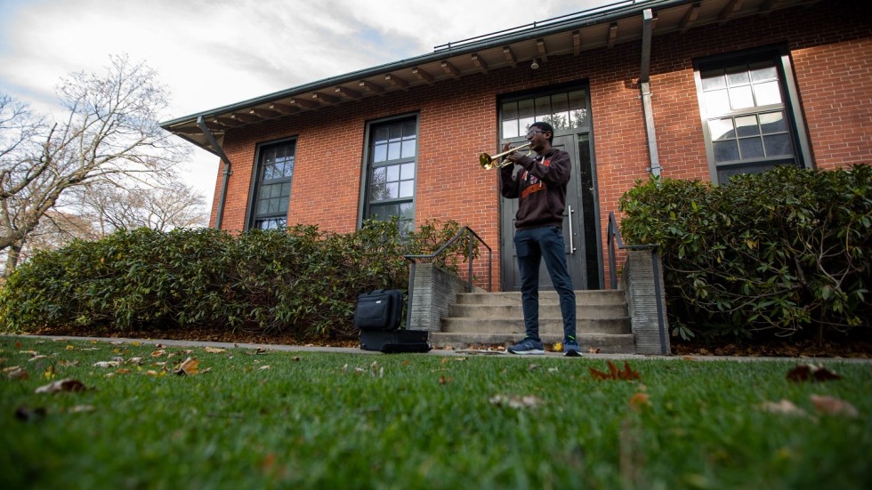 Derrick Pennix Jr. standing on grass, playing trumpet in front of a brick building