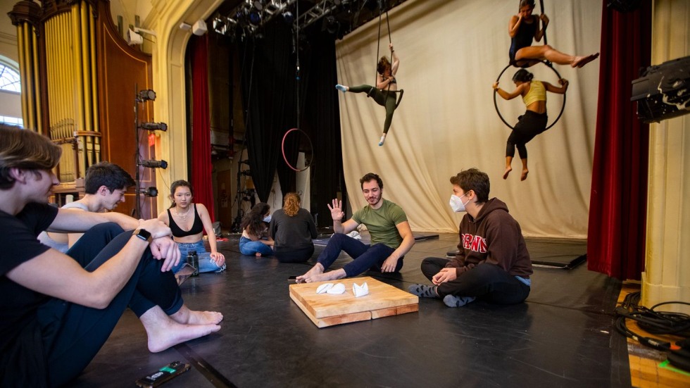students sitting and talking on a stage, with trapeze and lyra artists in the background